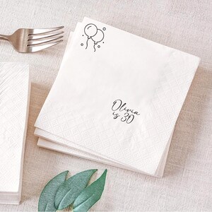 May include: White paper napkins with a diamond pattern and the text "Olivia is 30" in black script. Two black line-drawn balloons are in the upper right corner. A silver fork is visible in the upper left corner.
