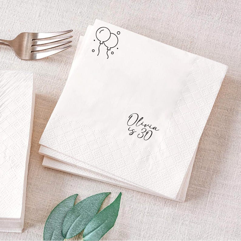 May include: White paper napkins with a diamond pattern and the text "Olivia is 30" in black script. Two black line-drawn balloons are in the upper right corner. A silver fork is visible in the upper left corner.