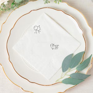 May include: White folded napkin with a black balloon graphic and the text "Olivia is 30". The napkin is placed on a white plate with a gold rim. Green leaves and floral accents are in the background.