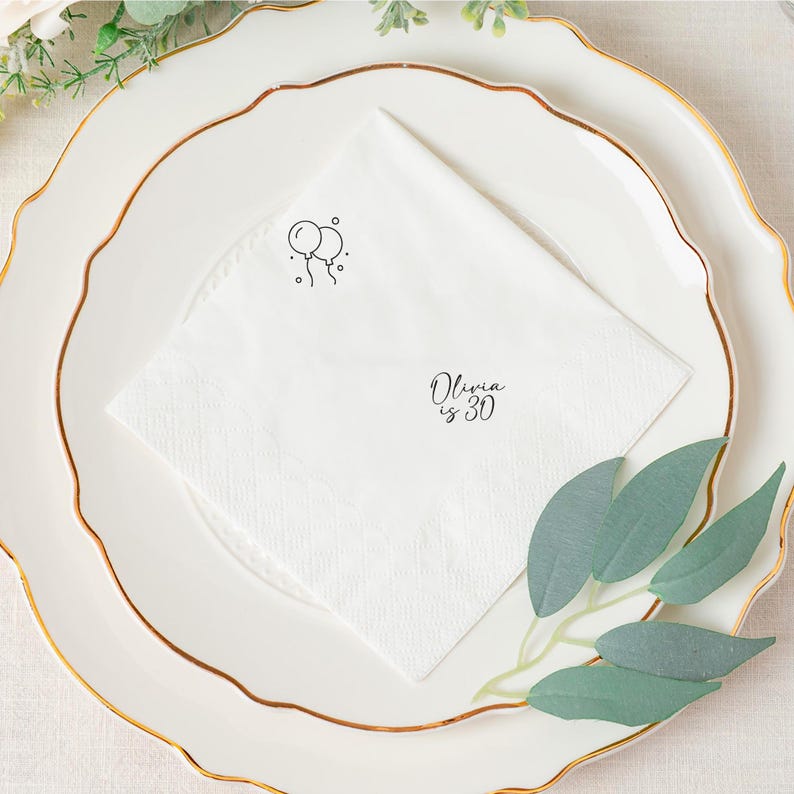 May include: White folded napkin with a black balloon graphic and the text "Olivia is 30". The napkin is placed on a white plate with a gold rim. Green leaves and floral accents are in the background.