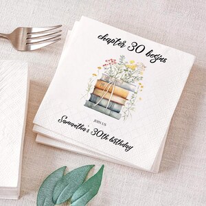 May include: A white square paper napkin featuring a stack of books with wildflowers on top. The text reads chapter 30 begins and JOIN US Samantha's 30th birthday in black script and serif fonts. A silver fork is visible in the upper left corner.