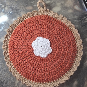 May include: Crocheted orange pot holder with a white, flower-shaped accent in the center. The pot holder has a tan-colored crust and a loop for hanging. The handmade kitchen item is round and resembles a pie.