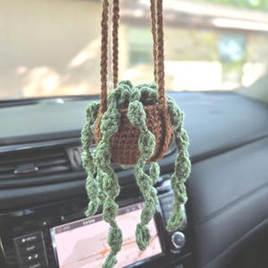 May include: A handmade crochet plant hanger with a brown pot and green, trailing plant. The plant hanger is suspended from a brown cord, designed to hang from a car's rearview mirror or similar location. The interior of a car is visible in the background.