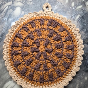 May include: A round, crocheted potholder in shades of brown and tan. The center features a dark brown floral design, surrounded by a lighter brown pattern. The edge is scalloped and tan, with a loop for hanging. A handmade kitchen accessory.