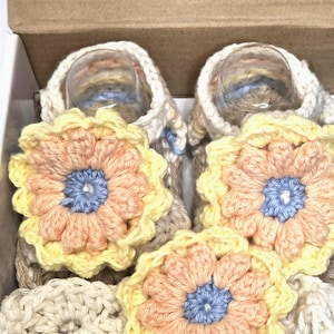 May include: Handmade crochet baby booties and matching flower appliques in a cardboard box. The booties are beige, with yellow, peach, and blue flower accents. The appliques are peach, yellow, and blue.