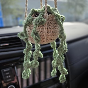 May include: A handmade crochet plant hanger with a beige pot and green, trailing plant. The plant hanger is suspended by beige rope, creating a charming, bohemian aesthetic. The plant hanger is perfect for adding a touch of nature to any space.