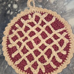 May include: A handmade, round crochet hot pad with a pie crust design. The center is a deep burgundy color, with a lattice pattern in a light tan color. The edge is scalloped and tan, with a loop for hanging.