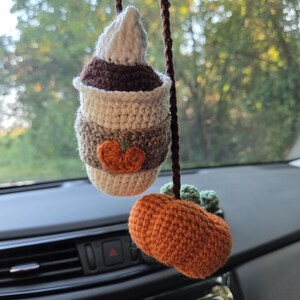 May include: Crocheted car charm featuring a coffee cup with whipped cream, a pumpkin, and green accents. The coffee cup is brown, tan, and white, with an orange pumpkin applique. The pumpkin is orange with green leaves.