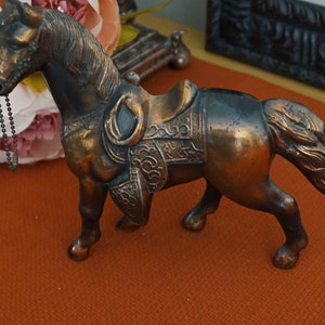 Könnte beinhalten: Eine bronzene Pferdefigur mit Sattel und einer Kette, die aus seinem Maul hängt. Das Pferd steht auf einer braunen Oberfläche.