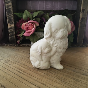May include: White ceramic figurine of two Pekingese dogs sitting together. The larger dog has its head turned to the side and the smaller dog is nestled against its side.