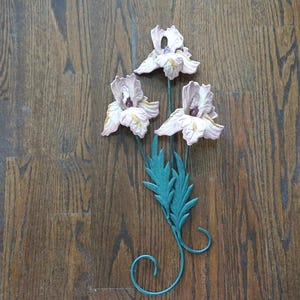 Op de afbeelding: Een decoratieve metalen wanddecoratie met drie irisbloemen in wit- en paarstinten. De bloemen zijn bevestigd aan een turquoise stengel met bladeren en een gekrulde basis. De achtergrond is een houten oppervlak.