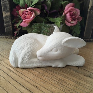 Peut inclure: Figurines en céramique blanche d'un faon couché, la tête reposant sur ses pattes. Le faon est sculpté avec des détails réalistes, y compris la fourrure et les yeux.