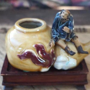 May include: A small, ceramic decorative piece featuring a yellow pot with a maroon design and a seated figurine. The figurine is dressed in blue and tan clothing. The piece is set on a small, dark wooden base.