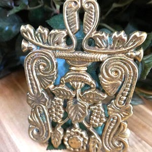 May include: A rectangular brass trivet featuring an intricate openwork design of embossed leaves, swirling scrolls, and clusters of grapes. The metal surface displays a polished gold-toned finish with subtle darkened patina in the recessed areas.