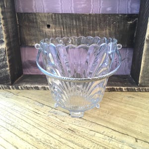 May include: A clear glass bucket with a scalloped rim and a vertical ribbed pattern. The bucket features a silver-toned metal handle with a textured finish and stands on three small glass feet, providing a stable base for the vessel.