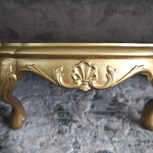 May include: A gold-colored display stand featuring a flat circular top and three curved legs. The apron of the stand is decorated with a central shell motif flanked by scrolling leaf patterns, providing a raised platform for small decorative items.