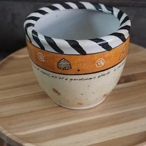 May include: A ceramic planter with a black and white striped rim, an orange band featuring small illustrations of a beehive and garden tools, and a cream-colored base with speckled details and cursive text that reads, all of a gardener's efforts.