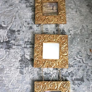 May include: Three ornate, gold-colored picture frames hanging vertically. The top frame has a decorative top piece. Each square frame is connected by a chain. The frames contain a landscape, a blank space, and a still life painting.