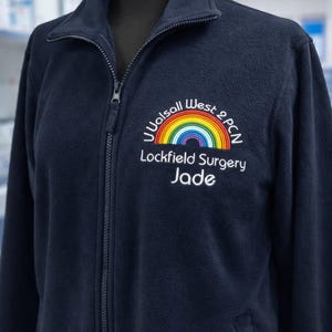 May include: Navy blue fleece jacket with a full zipper and a high collar. The jacket features an embroidered rainbow design with the text "Walsall West 2 PCN, Lockfield Surgery, Jade".
