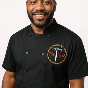 May include: A black chef's jacket with short sleeves and a mandarin collar. The jacket features a round embroidered logo that says "Kymari's TOP CHEF" with a knife graphic. The jacket has black buttons and is worn by a smiling person.