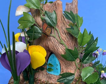 May include: A fantasy fairy house sculpture. A tree trunk with a blue door is surrounded by paper flowers in purple, yellow, and white. Green leaves and a butterfly add to the scene. Mushrooms and grass complete the miniature garden setting.