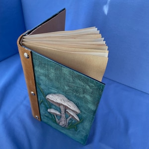 May include: A handmade journal with a leather spine and a teal cover featuring a 3D mushroom design. The journal's pages are a light tan color, and the spine is secured with silver rivets. The journal is open, revealing the blank pages.