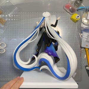 May include: A detailed figurine of a character with long white hair accented with blue streaks. The figure is dressed in a black and blue outfit and stands on a white base. The hair flows dramatically, creating a dynamic pose.