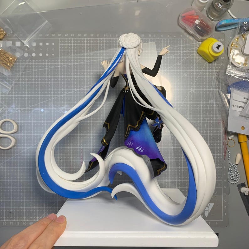 May include: A detailed figurine of a character with long white hair accented with blue streaks. The figure is dressed in a black and blue outfit and stands on a white base. The hair flows dramatically, creating a dynamic pose.