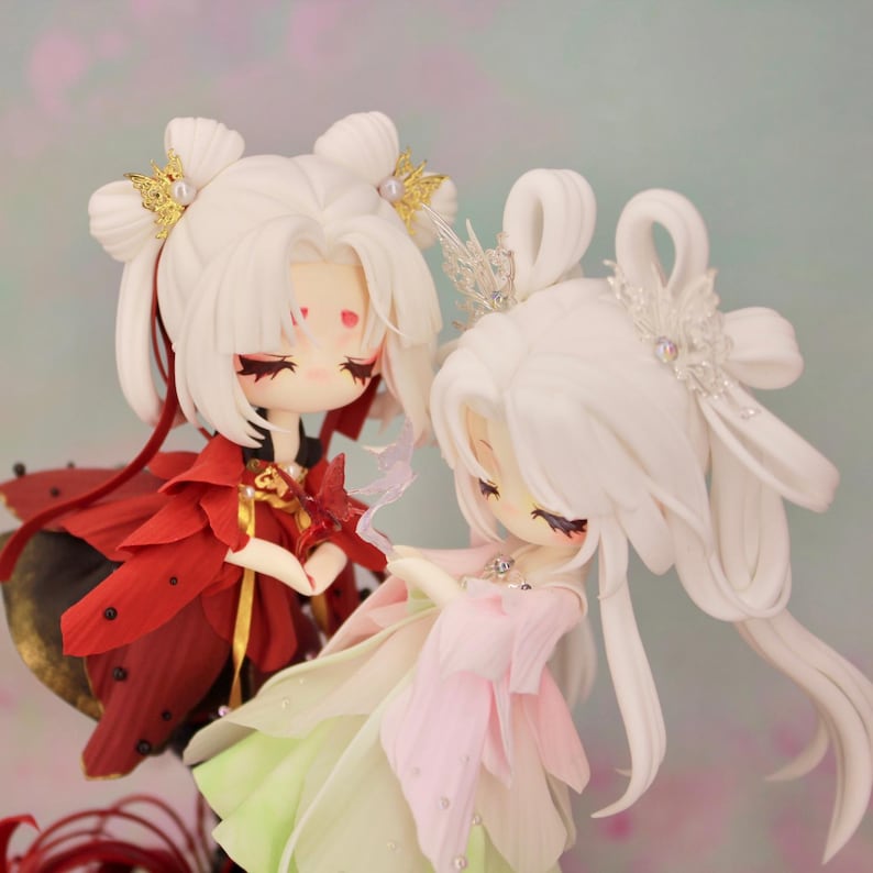 May include: Two detailed figurines of women with white hair and elaborate headpieces. One wears a red petal-like dress, the other a pink and green petal dress. Both have closed eyes and delicate facial features, with a soft, blurred background.