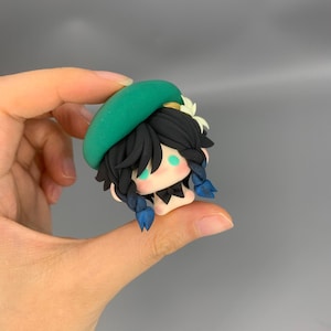 May include: A small, handcrafted figurine of a character with black hair, blue braids, and a green beret. The figurine has a white flower and a black bow tie. The character's face has light green eyes and a pale complexion.