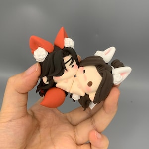 May include: Two small, handcrafted figurines in a kissing pose. One figurine has black hair, red fox ears, and a red tail. The other has brown hair and white bunny ears. Both have rosy cheeks and are made of clay.