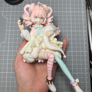 May include: A detailed figurine of a seated anime-style character with pink hair and blue eyes. The character wears a white dress with pastel accents, striped stockings, and holds a chocolate bar and a plushie. The figurine is held in a hand.