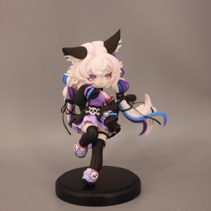 May include: A detailed figurine of a character with pink hair, black and white ears, and purple eyes. The figure wears a black and purple outfit with skull accents and black stockings. It stands on a black circular base.