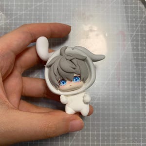 May include: A small, white figurine of a character wearing a bunny-eared hood. The character has gray hair, blue eyes, and a small, smiling face. The figurine is held against a gray cutting mat.