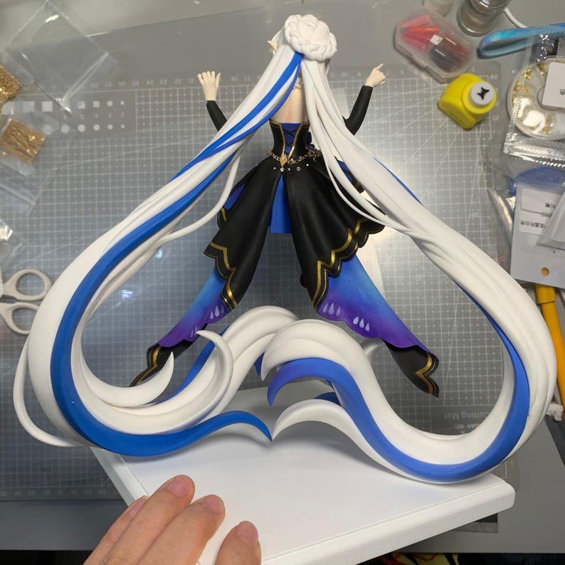May include: A detailed figurine of a character with long white hair, accented with blue streaks. The figure wears a black and blue dress with gold trim and is posed with arms raised. The base is white, and the hair forms a heart shape.