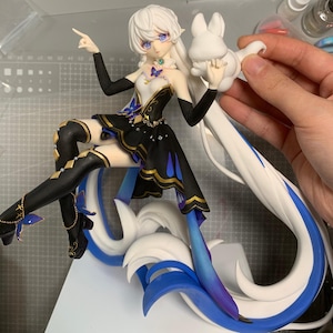 May include: A detailed figurine of a female anime character with white hair and blue eyes. She wears a black and gold dress with butterfly accents. The figure is posed with a rabbit-shaped accessory and sits atop a stylized base with blue and white accents.