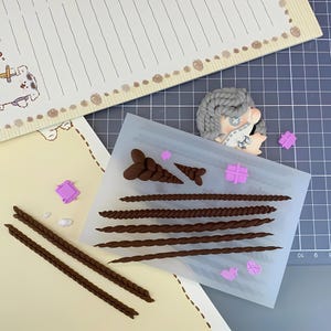 Puede incluir: Un proyecto de manualidades con un molde de silicona con varios diseños de trenzas y pequeñas formas rosas. También hay dos trenzas de arcilla marrón y una cabeza de muñeca de pelo gris. El fondo incluye papel rayado y una esterilla de corte.