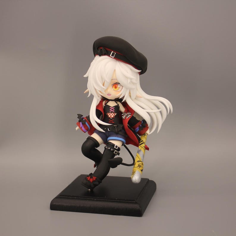 May include: A figurine of an anime-style character with white hair, wearing a black beret, red jacket, and black shorts. The figure is holding a yellow and silver object and is mounted on a black square base.