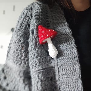 May include: A handmade crochet mushroom pin with a red cap and white polka dots is attached to a gray crochet shawl. The mushroom pin is a whimsical accessory, adding a touch of nature-inspired charm to the shawl. The shawl is made of a chunky, textured yarn.