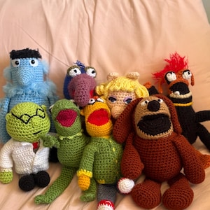 Peut inclure: Une collection de personnages des Muppets faits à la main au crochet, dont Kermit la Grenouille et le Dr. Bunsen Honeydew. Chaque personnage est fabriqué avec du fil de couleur vive, présentant des traits et des expressions détaillés. Les jouets sont disposés sur une surface douce de couleur pêche.