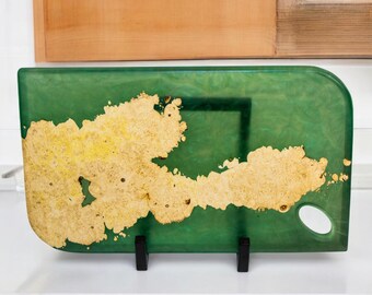 May include: A rectangular green resin cutting board with a natural wood grain design. The board has rounded corners and a small, circular hole. The wood grain is a light tan color and is spread across the surface.