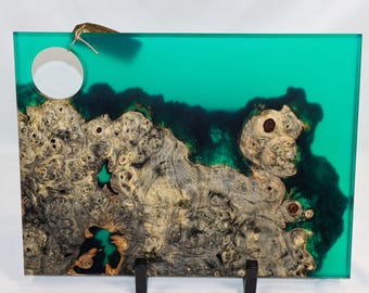 May include: Rectangular wooden serving board with a teal resin top. The wood grain is visible, with a natural, organic pattern. The board has a hole in the upper left corner and is supported by two black stands. A small tag is attached.