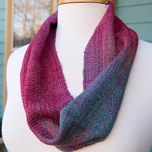 May include: A handwoven infinity scarf with a two-tone design. One side is a deep burgundy color with a subtle shimmer, while the other side is a teal blue with a similar shimmer.