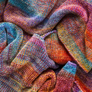 May include: A close-up of a colorful woven fabric with a textured surface. The fabric is a blend of orange, red, blue, and purple hues.