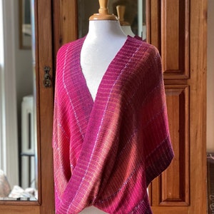 May include: A pink and orange woven shawl with a diagonal pattern. The shawl is draped over a white mannequin.