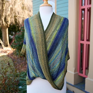 May include: A knitted shawl in shades of blue, green, and olive green is draped over a white mannequin. The shawl features a V-neck design and a striped pattern. The background includes a house with blue siding and pink trim, and a tree.