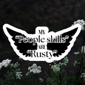 May include: A white sticker with black text and wing-shaped design. The text reads "MY 'People skills' ARE 'Rusty'". The sticker is set against a blurred background of dark green foliage and white flowers.