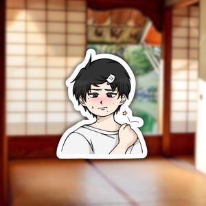 May include: A sticker featuring an anime-style illustration of a young person with black hair, a white t-shirt, and a bandage on their head. The character has a blush and a star graphic near their shoulder.