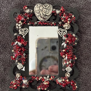 May include: A small, rectangular black mirror with ornate detailing. The frame is embellished with red and black beads, silver hearts, and a silver butterfly. A silver heart with a floral design is at the top.