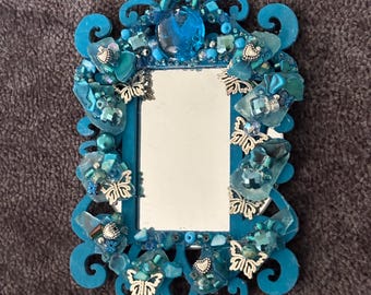 May include: A small, rectangular mirror with a vibrant turquoise frame. The frame is adorned with various shades of blue beads, crystals, and silver butterfly accents. The mirror is a decorative item.